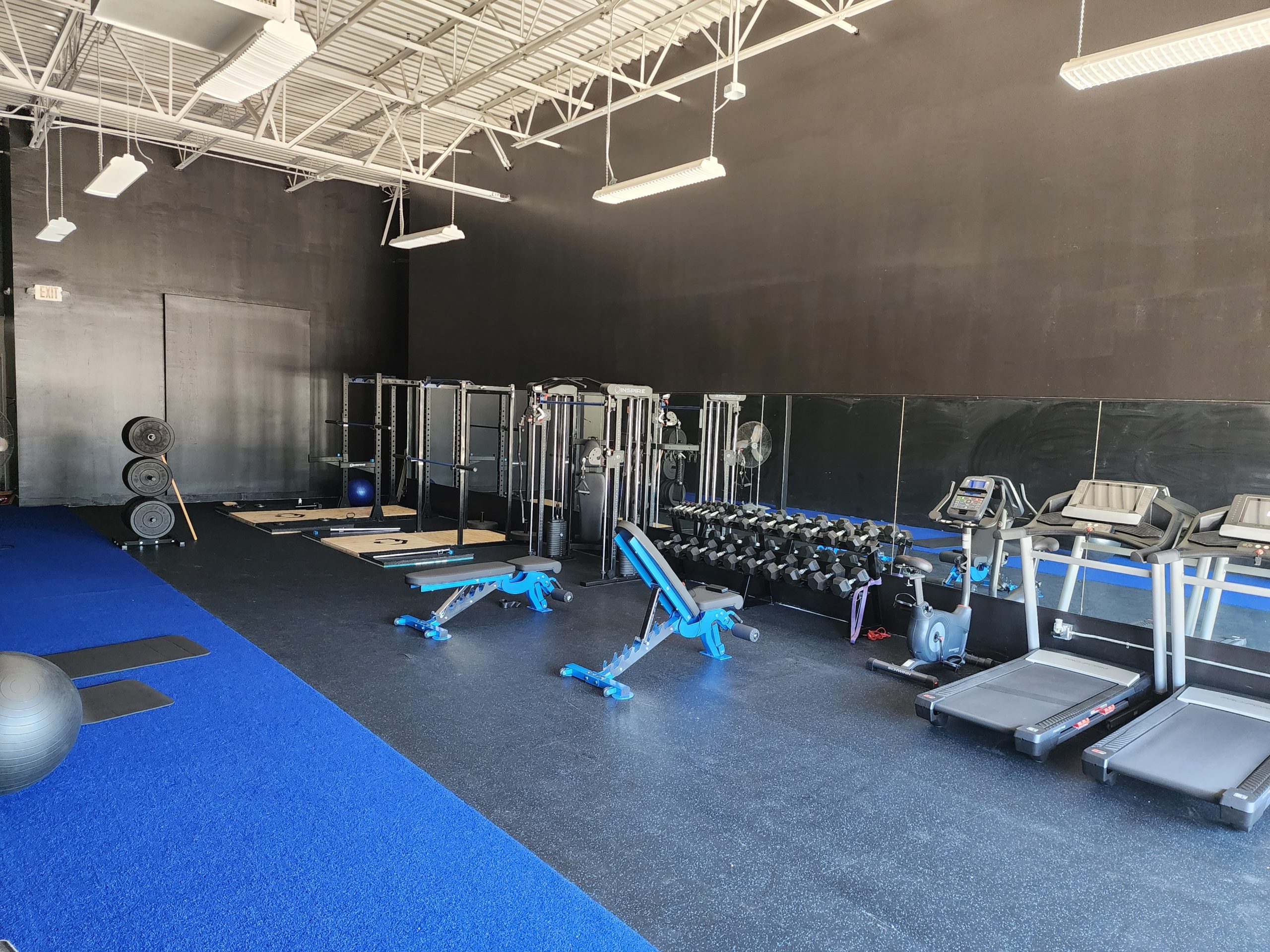 about us- the inside of the Fitness Minded personal training studio which shows from right to left 2 treadmills, 2 benches, dumbbell set, 2 squat racks, and blue turf